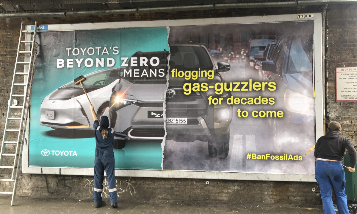 A billboard shows, on the left, a Toyota advert touting their sustainability, and, on the right, a traffic jam of heavily polluting cars, one of which has a Toyota logo very prominently on its front.