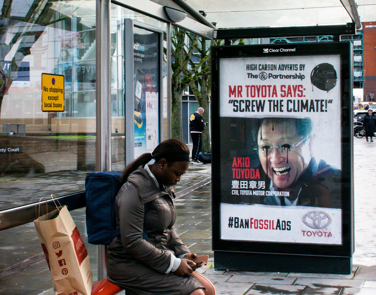 A poster shows a man's face with oil pouring from the eyes. A caption reads: "Mr Toyota says Screw the Climate"