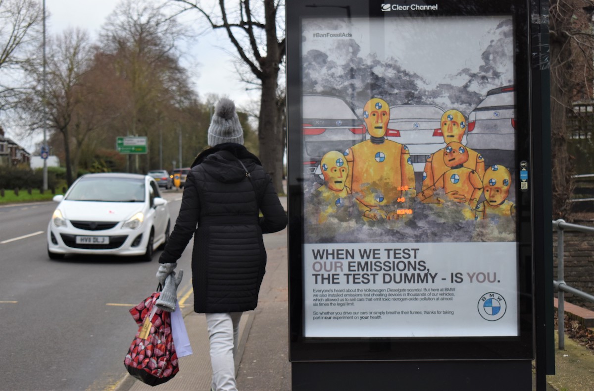 A poster in a bus stop shows crash test dummies, made to look like a family of two parents and two children, above the caption "When we test our emissions, the test dummy is you"