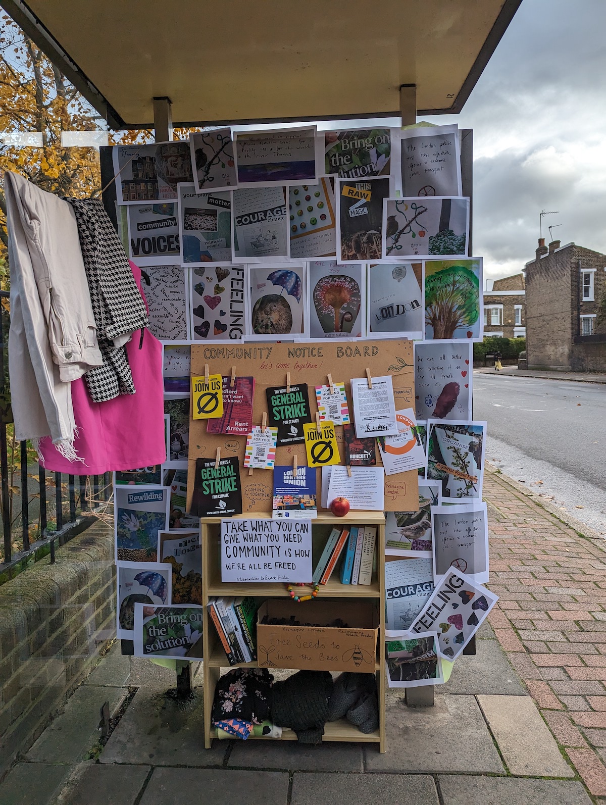 a bus stop covered with printed images of different things. In the middle on the ground is a bookcase with a sign reading "Take what you can. Give what you need. Community is how we're all be freed"