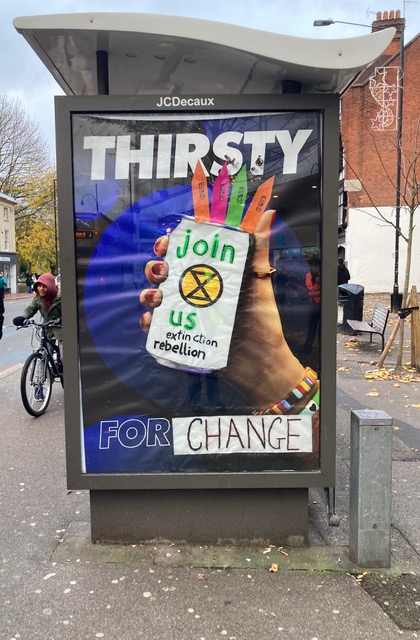 A bus stop poster shows a hand holding a can bearing the XR logo and the words "join us". Around the can are the words "thirsty for change"